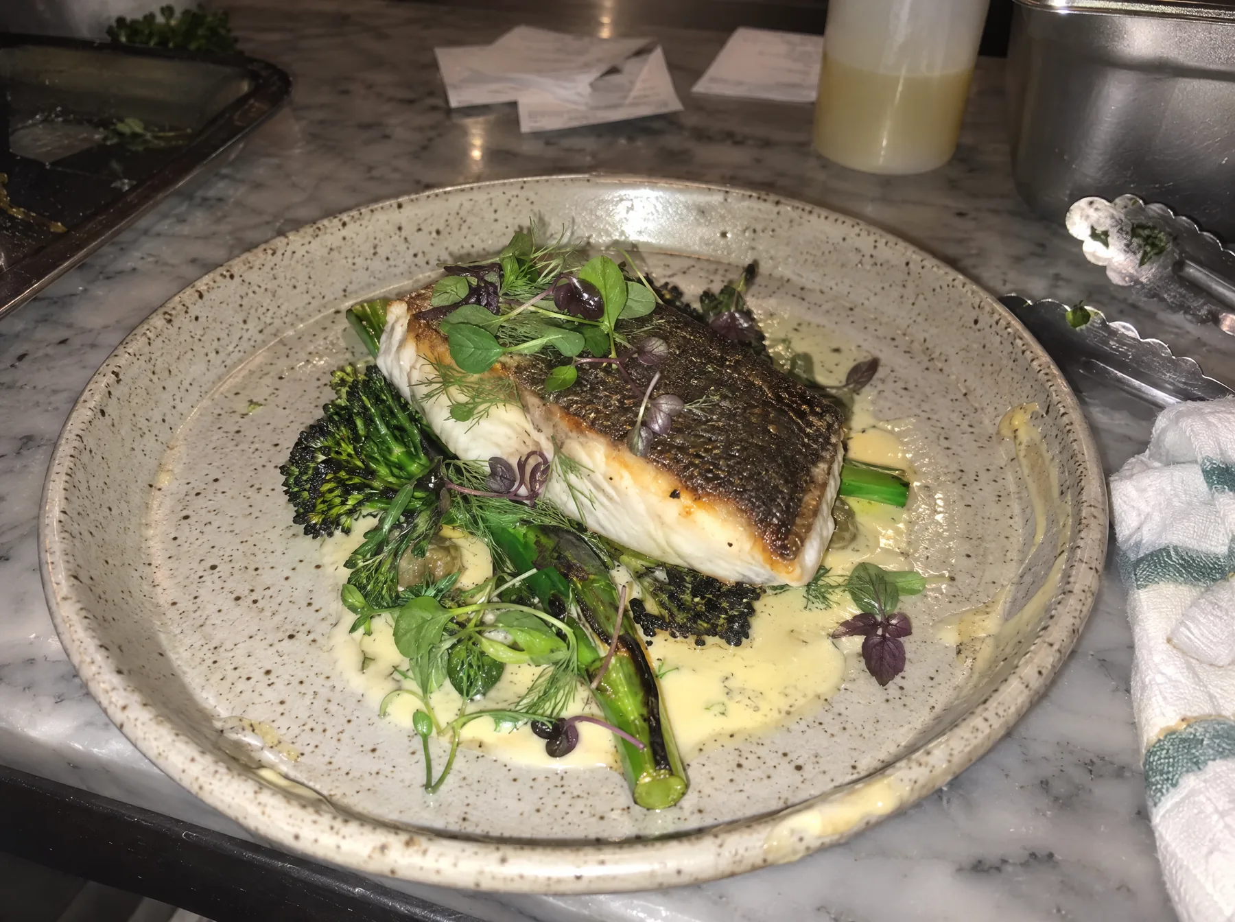 Before: barramundi on a speckled plate under harsh flash, with tickets and tongs on the pass