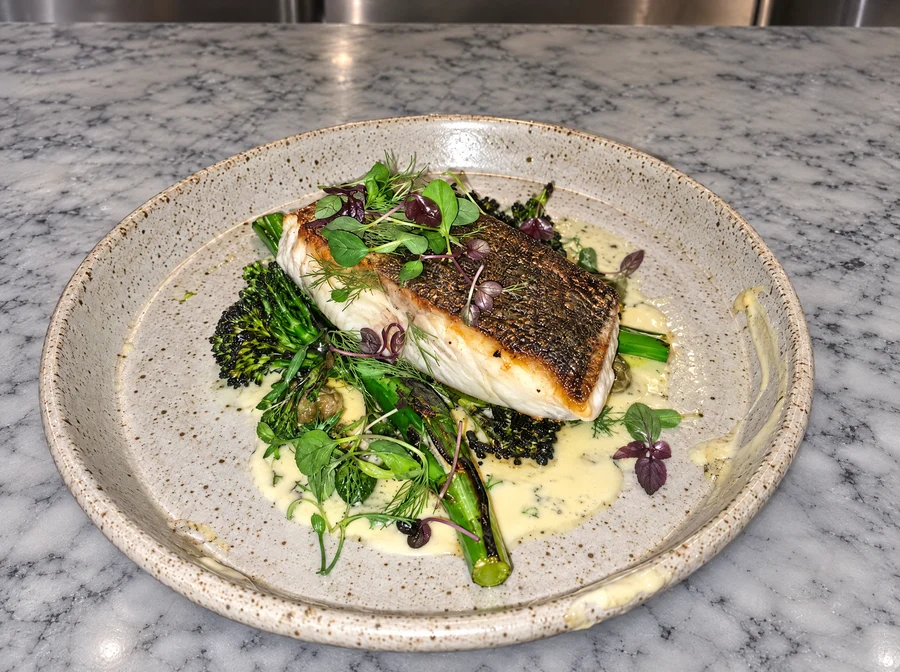 Pan-roasted fish fillet with crisp skin on charred broccolini and beurre blanc, in a speckled stoneware bowl on white marble